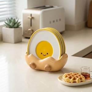 Könnte beinhalten: Ein Set aus vier runden Keramiktellern mit einem gelben Eigelb-Design und einem kleinen lächelnden Gesicht, die aufrecht in einem hellbeigen, eiförmigen Keramikständer mit zwei kleinen abgerundeten Füßen an der Basis stehen.
