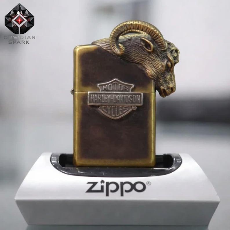 Puede incluir: Encendedor Zippo de color lat&oacute;n con una cabeza de carnero y un emblema de Harley-Davidson. El encendedor est&aacute; sobre un soporte Zippo blanco. El logotipo "Obsidian Spark" es visible. Mide unos 5,5 cm de alto.
