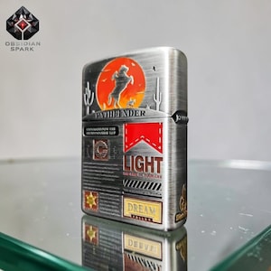 May include: A silver-toned metal lighter with a desert-themed design. The lighter features a silhouette of a horse and rider against an orange sunset, with the word "PATHFINDER" above. It also has the word "LIGHT" and the phrase "THE FIRE OF YOUR LIFE".