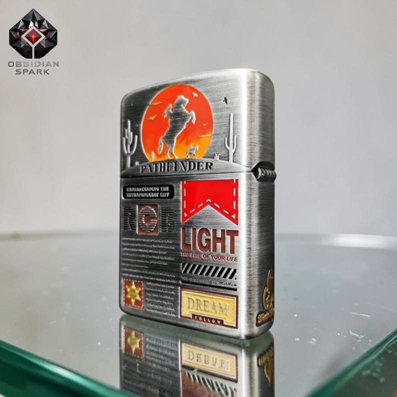 May include: A silver-toned metal lighter with a desert-themed design. The lighter features a silhouette of a horse and rider against an orange sunset, with the word "PATHFINDER" above. It also has the word "LIGHT" and the phrase "THE FIRE OF YOUR LIFE".