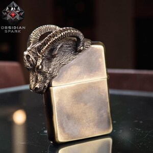Puede incluir: Un encendedor de color bronce con un dise&ntilde;o detallado de cabeza de carnero en la parte superior. El encendedor tiene forma rectangular y un acabado met&aacute;lico. La marca "OBSIDIAN SPARK" es visible.