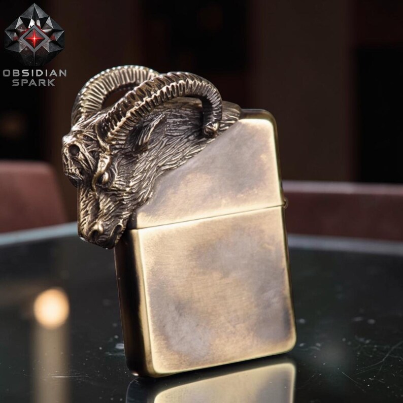 Puede incluir: Un encendedor de color bronce con un dise&ntilde;o detallado de cabeza de carnero en la parte superior. El encendedor tiene forma rectangular y un acabado met&aacute;lico. La marca "OBSIDIAN SPARK" es visible.