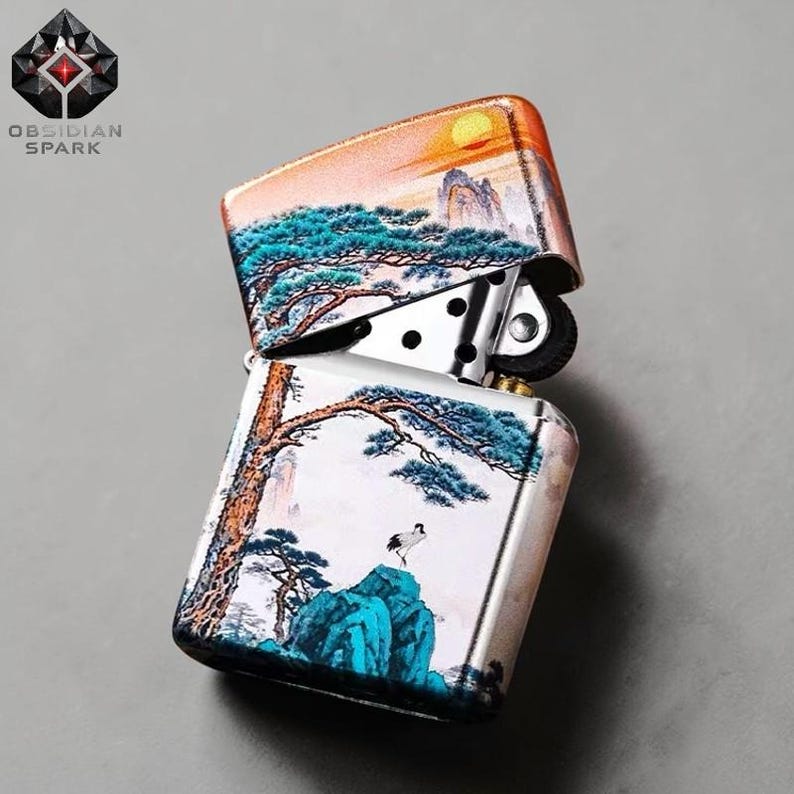 May include: A silver metal lighter with a landscape design. The top of the lighter features a sunset over mountains and trees. The bottom half shows a tree, a bird, and blue mountains. The brand name "OBSIDIAN SPARK" is visible.