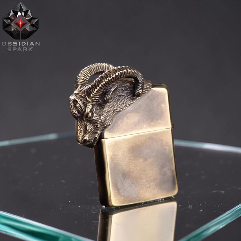 Puede incluir: Un encendedor de color lat&oacute;n con un dise&ntilde;o detallado de cabeza de carnero en la parte superior. El encendedor tiene forma rectangular y una tapa con bisagras. La marca "OBSIDIAN SPARK" es visible en la esquina superior izquierda.