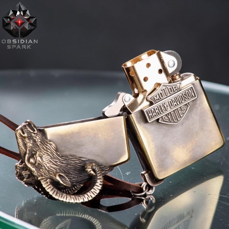 Puede incluir: Un encendedor de estilo vintage con acabado de lat&oacute;n, con el logotipo de Harley-Davidson y un dise&ntilde;o detallado de cabeza de carnero. El encendedor est&aacute; abierto, revelando su interior, y est&aacute; conectado a una correa de cuero. La marca "Obsidian Spark" es visible.