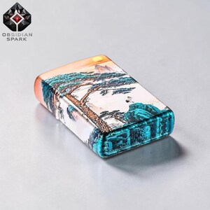 May include: A rectangular lighter with a landscape design featuring a tree, mountains, and a sunset. The lighter has a teal and orange trim and the text "OBSIDIAN SPARK" is visible.