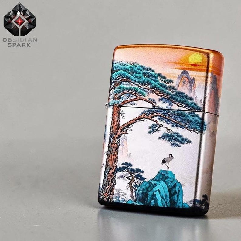 May include: A metal lighter with a scenic landscape design. The artwork features a large tree, mountains, and a crane. The top of the lighter has an orange and yellow gradient, and the brand name "OBSIDIAN SPARK" is visible.