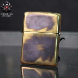 Puede incluir: Un encendedor de estilo vintage con acabado en lat&oacute;n y una p&aacute;tina &uacute;nica. El encendedor rectangular tiene bordes redondeados y una tapa con bisagras. La superficie muestra una mezcla de tonos dorados y morados, con el texto "OBSIDIAN SPARK" y un logotipo.