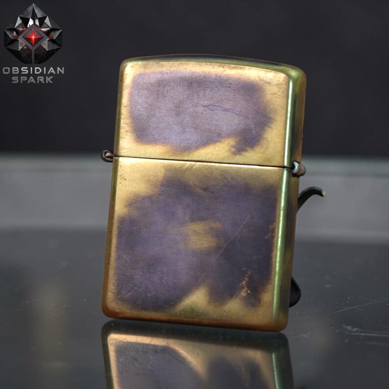 Puede incluir: Un encendedor de estilo vintage con acabado en lat&oacute;n y una p&aacute;tina &uacute;nica. El encendedor rectangular tiene bordes redondeados y una tapa con bisagras. La superficie muestra una mezcla de tonos dorados y morados, con el texto "OBSIDIAN SPARK" y un logotipo.