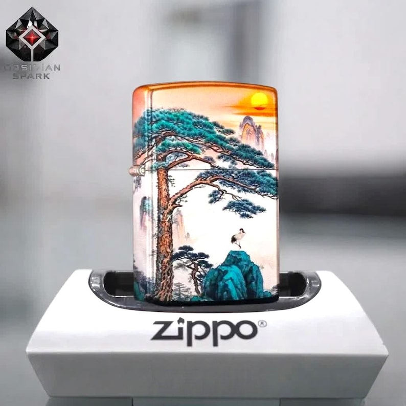 May include: A Zippo lighter with a scenic landscape design featuring a tree, mountains, and a sunset. The lighter is displayed on a white stand. The Zippo logo is visible on the stand.