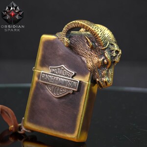 Puede incluir: Un encendedor de color bronce con un dise&ntilde;o de cabeza de carnero en la parte superior. El encendedor presenta el logotipo de Harley-Davidson y el texto "OBSIDIAN SPARK". El encendedor tiene una correa de cuero.