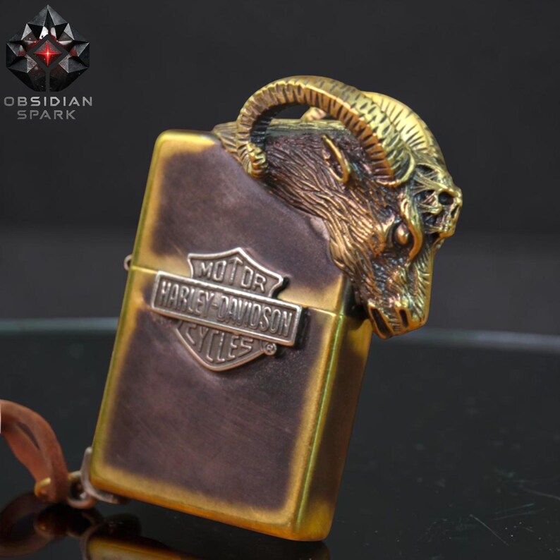 Puede incluir: Un encendedor de color bronce con un dise&ntilde;o de cabeza de carnero en la parte superior. El encendedor presenta el logotipo de Harley-Davidson y el texto "OBSIDIAN SPARK". El encendedor tiene una correa de cuero.