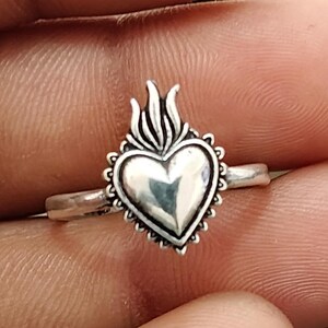 Può includere: Un anello color argento con un emblema centrale a forma di cuore sormontato da un motivo a fiamma. Il cuore è circondato da un bordo decorativo smerlato con piccoli accenti circolari, montato su una fascia in metallo color argento liscia e lucida.