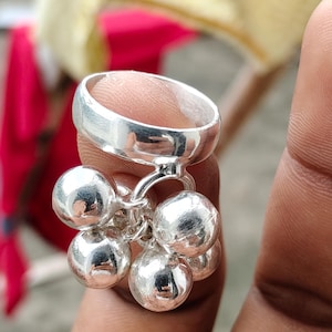 May include: A polished silver ring featuring a cluster of five dangling spherical charms. The band is smooth and circular, while the attached charms are round, reflective, and metallic, creating a cohesive silver-toned accessory design.