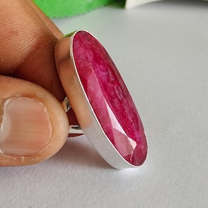Peut inclure: Une bague ornée d'une grande pierre précieuse rubis facettée de forme ovale allongée, sertie dans une monture en argent lisse et polie. La pierre rouge rosée profonde présente une surface texturée à facettes qui capte la lumière.