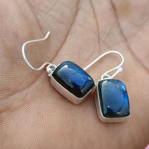 Puede incluir: Un par de pendientes colgantes de plata con piedras preciosas de labradorita rectangulares que presentan un brillo iridiscente azul profundo. Cada piedra está engastada en un marco de plata liso con un gancho sencillo.
