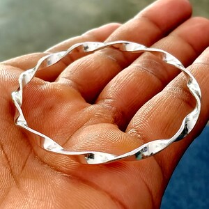 Può includere: Un bracciale rigido color argento lucido caratterizzato da un design continuo intrecciato e ondulato. La superficie metallica è liscia e riflettente, creando una serie di curve ripetitive che formano una forma circolare di spessore uniforme.