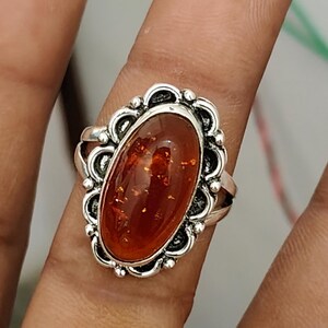 May include: An oval-shaped amber gemstone set in a decorative silver ring. The amber is a deep, translucent orange color with visible internal inclusions. The silver setting features a scalloped, ornate border surrounding the central stone.