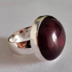 Può includere: Un anello in argento con una grande pietra cabochon rotonda, liscia e rosso scuro incastonata in una fascia di metallo lucido. L'interno della fascia in argento è chiaramente marchiato con il punzone 925, che indica la composizione del metallo.