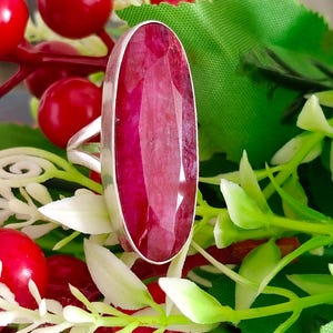 Pode incluir: Um anel de prata com uma grande pedra preciosa de rubi facetada de forma oval alongada, engastada numa moldura de metal lisa. A pedra vermelho-profundo apresenta uma superfície texturizada e multifacetada que capta a luz.