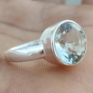 Puede incluir: Un anillo de plata con una gran piedra preciosa redonda y facetada de color verde pálido engastada en un bisel liso. La banda es de plata pulida con un diseño sencillo y sólido que se estrecha ligeramente hacia la parte posterior del anillo.