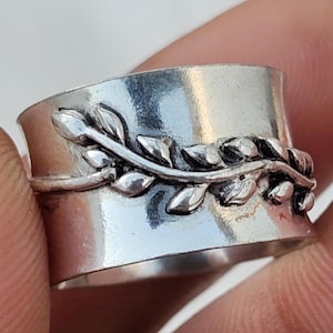 May include: A wide silver band ring featuring a raised, polished vine motif that wraps horizontally around the center. The metal has a smooth, reflective finish with darkened recessed areas that highlight the detailed leaf and stem design.