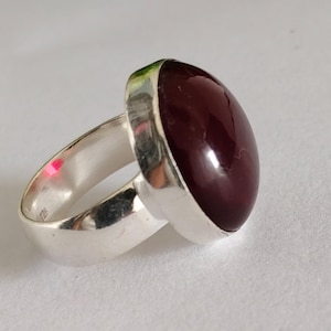 May include: A silver ring featuring a large, smooth, oval-shaped dark burgundy gemstone set in a polished metal bezel. The band is wide and solid, with a reflective metallic finish that catches the light, showcasing a simple and clean design.