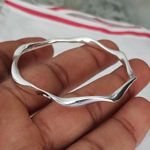 May include: A polished silver bangle bracelet featuring a continuous wavy, undulating design. The metal surface is smooth and reflective, creating a series of gentle curves that form the circular shape of the jewelry piece, which is held in place.