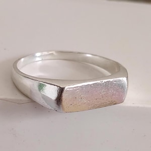 Könnte beinhalten: Ein silberfarbener Ring mit einer flachen, rechteckigen Vorderseite, die eine leicht texturierte, schillernde Oberfläche aufweist. Die Ringschiene ist glatt und poliert und verjüngt sich nach vorne hin zum rechteckigen Mittelstück. Die Metalloberfläche reflektiert das Licht.