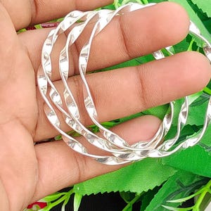 Può includere: Un set di tre braccialetti rigidi color argento caratterizzati da un design a spirale ritorto continuo. Ogni bracciale presenta una superficie lucida e riflettente che cattura la luce, creando una serie di curve ripetitive lungo gli anelli.