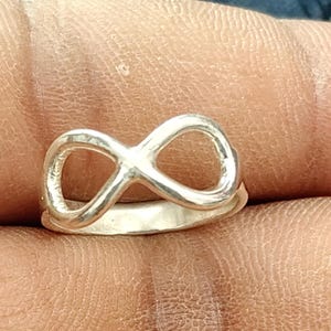 May include: A silver-toned ring featuring a prominent infinity symbol design on the front. The band is smooth and circular, connecting to the base of the infinity loop. The metal has a polished, reflective finish that catches the light.