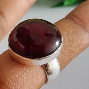 Op de afbeelding: Een ring met een grote, gladde, ronde cabochon edelsteen in diep donkerrood, gezet in een gepolijste zilverkleurige metalen band. De steen heeft een glanzend, reflecterend oppervlak en zit stevig in een eenvoudige metalen zetting.