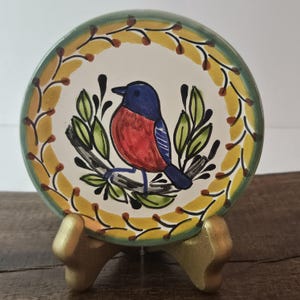 May include: A hand-painted ceramic plate featuring a colorful bird perched on a branch. The bird has a blue head, red chest, and blue tail feathers. The plate has a green rim and a yellow and brown floral border. The plate is displayed on a gold-colored stand.