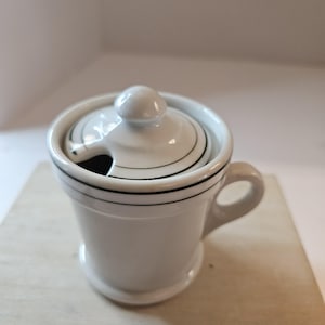May include: A white ceramic sugar bowl with a lid and a small handle. The bowl has a black stripe around the top and bottom. The lid has a knob and a slot for a spoon.