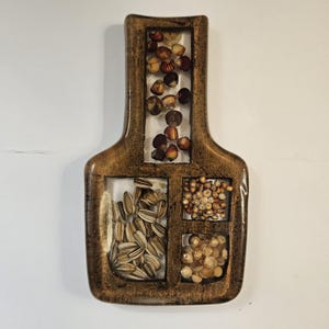 May include: A decorative wooden plaque with a distinctive design. The plaque is divided into sections, each filled with different types of seeds and nuts. The wooden frame has a warm, brown tone, and the seeds and nuts are in various shades of brown and tan.