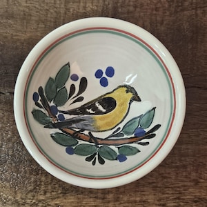 May include: A small, round ceramic bowl with a hand-painted design. The bowl features a yellow bird perched on a branch with green leaves and blue berries. The rim has red and green stripes. The bowl is white.