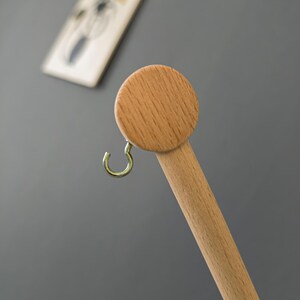 May include: A wooden coat rack with a round top and a gold-colored hook. The rack is made of light-colored wood and is set against a gray background. A framed picture with a yellow umbrella is in the background.
