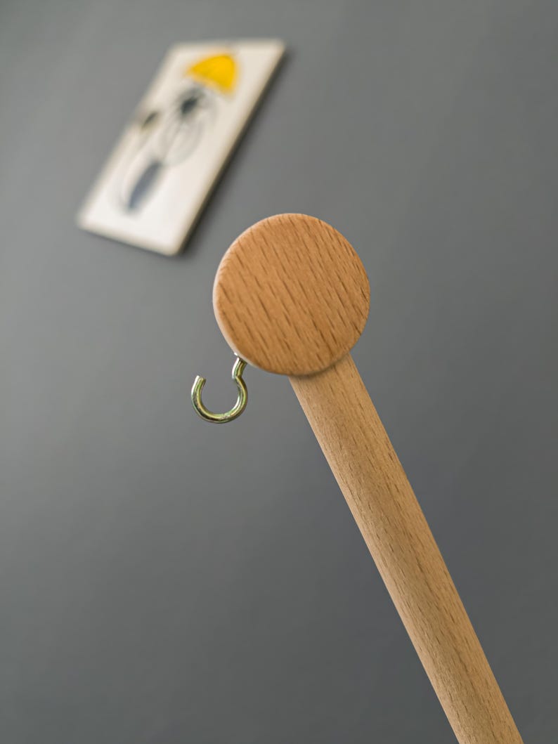 May include: A wooden coat rack with a round top and a gold-colored hook. The rack is made of light-colored wood and is set against a gray background. A framed picture with a yellow umbrella is in the background.