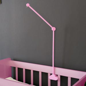 May include: A pink baby crib with vertical slats and a matching pink mobile arm. The mobile arm extends from the wall and has a curved design. The crib is set against a dark gray wall.