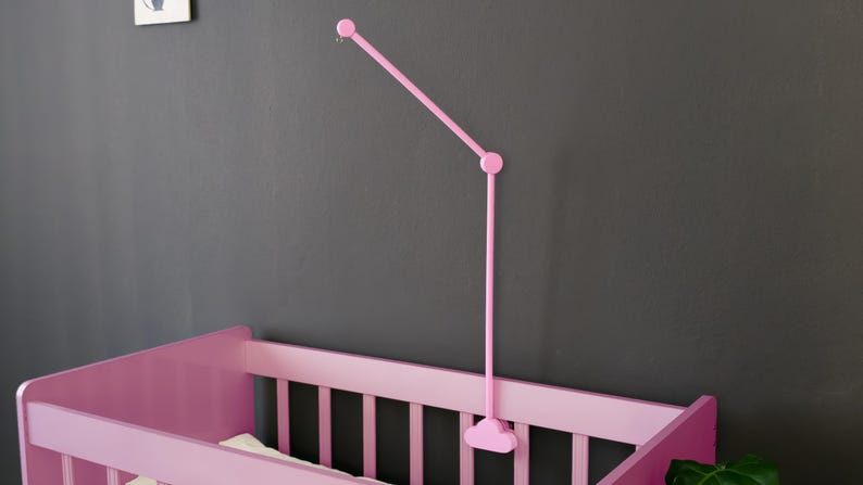 May include: A pink baby crib with vertical slats and a matching pink mobile arm. The mobile arm extends from the wall and has a curved design. The crib is set against a dark gray wall.