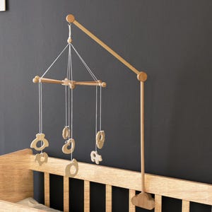 May include: Wooden baby mobile with hanging shapes, attached to a wooden crib. The mobile features a series of light-colored wooden cutouts suspended from a wooden frame. The crib is made of light-colored wood.