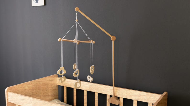 May include: Wooden baby mobile with hanging shapes, attached to a wooden crib. The mobile features a series of light-colored wooden cutouts suspended from a wooden frame. The crib is made of light-colored wood.