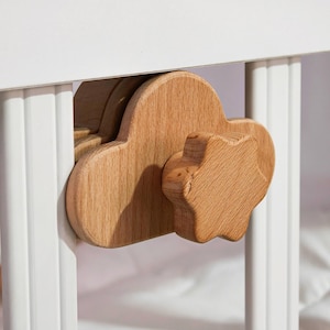May include: Close-up of a wooden cloud-shaped knob and a star-shaped knob attached to a white crib. The knobs are made of light-colored wood and are designed for easy gripping and turning. The crib is white.