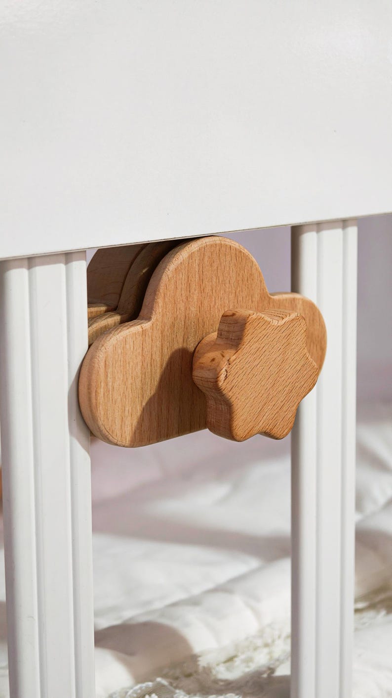 May include: Close-up of a wooden cloud-shaped knob and a star-shaped knob attached to a white crib. The knobs are made of light-colored wood and are designed for easy gripping and turning. The crib is white.