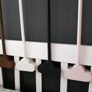 May include: A collection of cloud-shaped wooden mobiles in various colors, including natural wood tones, white, black, pink, and green. The mobiles are attached to long, slender rods and are displayed against a white crib railing and a dark gray wall.