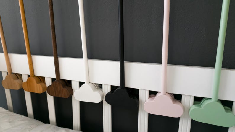 May include: A collection of cloud-shaped wooden mobiles in various colors, including natural wood tones, white, black, pink, and green. The mobiles are attached to long, slender rods and are displayed against a white crib railing and a dark gray wall.