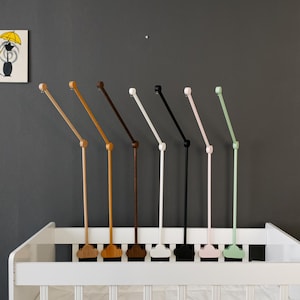 May include: A collection of baby mobile arms in various colors, including natural wood, brown, black, white, pink, and green, are displayed above a white crib. A small framed picture of a cat with an umbrella hangs on the gray wall.