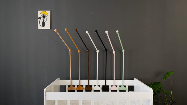 May include: A collection of baby mobile arms in various colors, including natural wood, brown, black, white, pink, and green, are displayed above a white crib. A small framed picture of a cat with an umbrella hangs on the gray wall.