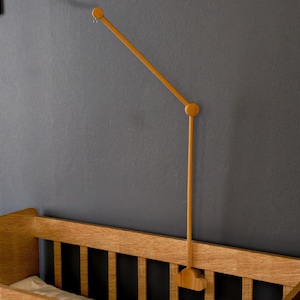 May include: Wooden baby crib with a matching wooden mobile arm attached to the wall. The crib is made of light-colored wood and has a simple, classic design. The mobile arm is a light brown color and extends over the crib.