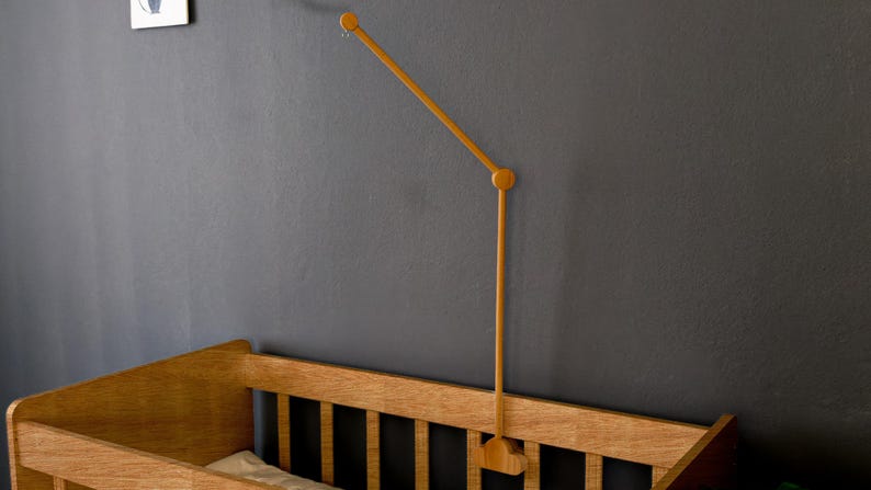 May include: Wooden baby crib with a matching wooden mobile arm attached to the wall. The crib is made of light-colored wood and has a simple, classic design. The mobile arm is a light brown color and extends over the crib.